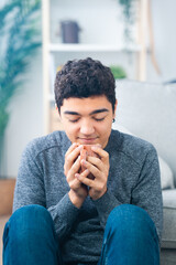 Fototapeta na wymiar Hispanic teenager boy sitting on floor smelling cup of coffee.