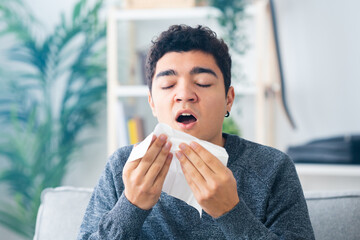 Hispanic teenager boy sneezing on tissue indoors. Illness and virus concept