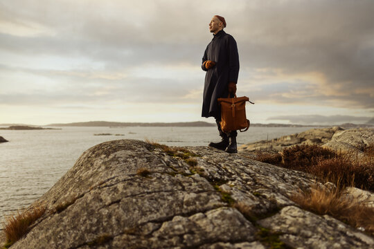 A Caucasian Man Wearing A Blue Coat And Holding A Backpack Standing On Top Of A Cliff By The Coast Looking Out Over The Ocean.