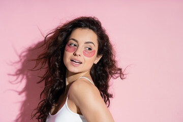 Curly young woman with hydrogel eye patches looking away on pink background.