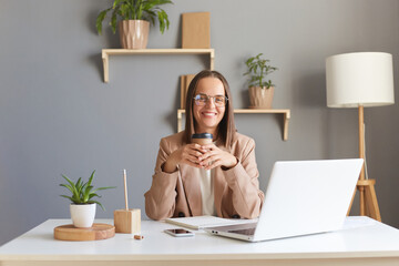 Photo of smiling joyful cheerful Caucasian woman wearing beige jacket posing in office, sitting at...