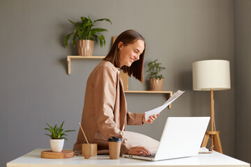 Side view portrait of smiling positive Caucasian woman wearing beige jacket posing in office, typing on notebook, holding paper documents, working at her workplace.