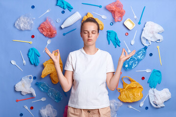 Fototapeta Portrait of calm woman dressed in white T-shirt, picked plastic litter isolated on blue wall, practicing yoga, trying to calm down, worries about the environment and causing harm to nature. obraz