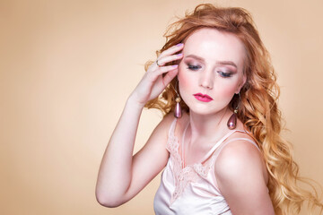 Beautiful red-haired girl with wavy hair, make-up, in a silk top and long earrings posing on a beige background in the studio. Woman touching her face and looking down