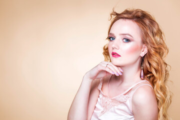 Fototapeta premium Beautiful red-haired girl with wavy hair, make-up, in a silk top and long earrings posing on a beige background in the studio. Woman touching her face and looking at the camera