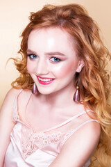 Naklejka premium Beautiful red-haired girl with wavy hair, make-up, in a silk top and long earrings posing on a beige background in the studio. Woman looking at the camera and smiling