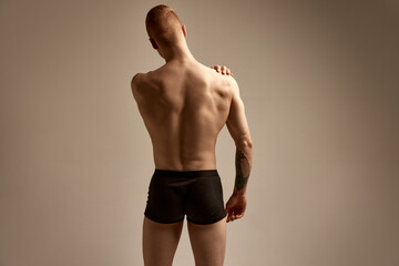 Fototapeta na wymiar Back view. Young redhead boy posing in black boxers, underwear over beige studio background. Strong, relief body shape. Concept of men's health and beauty, body and skin care, fitness. Body art