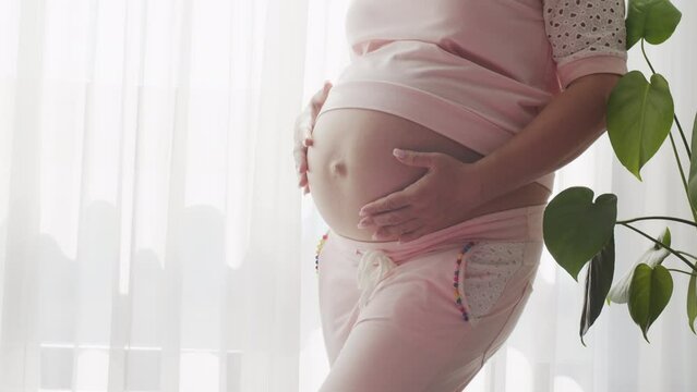 Pregnant woman expecting a baby. Adult pregnant woman feeling happy at home while strokes her child in  a belly. Pregnant woman  near window at home strokes touches her big belly. Slow motion