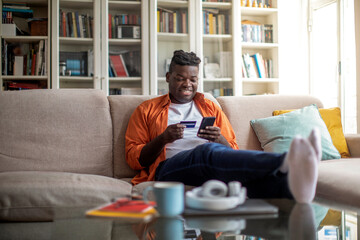 Cheerful african man using phone and credit card at home