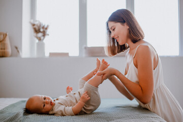 Caring young mother kissing baby's feet while making gymnastics on bed at home, side view, copy...