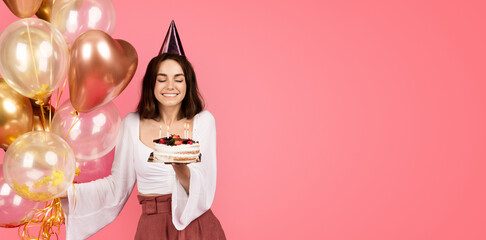 Cheerful young caucasian woman in hat hold a lot of inflatable balloons, blows out candles on cake