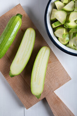 One green zucchini with two section halves on a cutting board with chopped zucchini in a bowl on a wooden background.