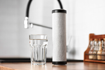 A glass of clean fresh water and carbon filter cartridge on wooden table in a kitchen interior. Installation of reverse osmosis water purification system. Concept Household filtration system. - obrazy, fototapety, plakaty