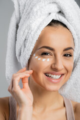 Positive woman with towel on head and cream on face looking at camera isolated on grey.