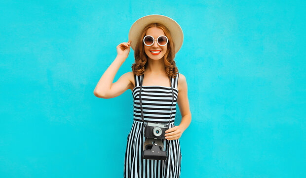 Summer portrait of happy smiling young woman photographer with vintage film camera wearing straw hat, dress, sunglasses on blue background