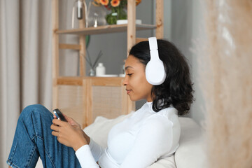 Side view portrait of a happy african amercian girl listening music,podcast or lecture on sofa at home with phone in her hands. Online application and modern technologies concept.