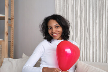 Closeup portrait of Young african american woman holding red heart shaped balloon sitting on sofa at home. Celebrating valentines,love day, women's day, birthday. Copy space banner