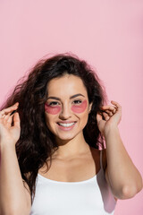 Cheerful young woman with hydrogel patches touching hair on pink background.