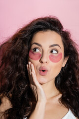 Dreamy woman with eye patches on face touching cheek on pink background.