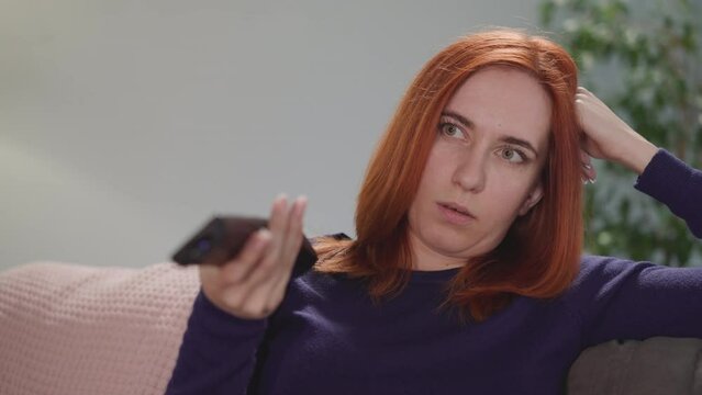 Distressed woman tries to find interesting TV program to watch at home after work. Redhead housewife leans head on hand holding remote control closeup