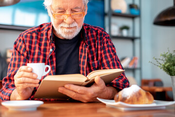 Senior bearded man sitting at cafe table with a book while enjoying reading pleasure and breakfast with espresso coffee cup and croissant. Elderly caucasian male in eyeglasses and checkered shirt