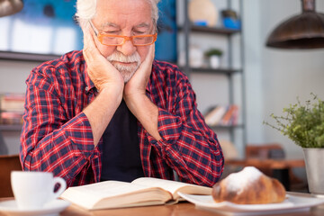 Serious senior bearded man sitting at cafe table reading a book while enjoys coffee cup and croissant. Elderly caucasian male in checkered shirt with eyeglasses