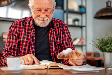 Handsome senior bearded man sitting at cafe table reading a book while enjoying breakfast with espresso coffee cup and croissant. Elderly caucasian male in eyeglasses and checkered shirt