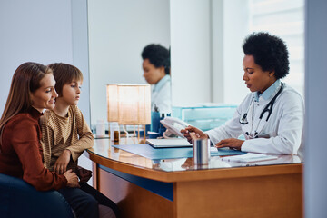 African American female doctor talks to mother and son during medical appointment at pediatrician's office.