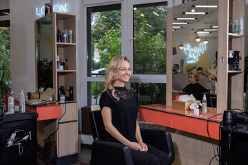 Blonde woman in black blouse sits on chair near workplace of makeup artist with large mirror. Person smiles looking contentedly in modern beauty salon