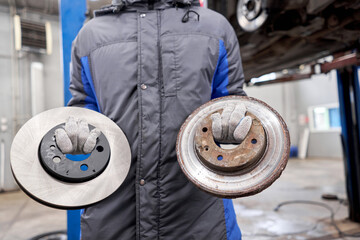 The mechanic holds old rusty brake disc and new disc. Change the old to new brake disc on car in a...