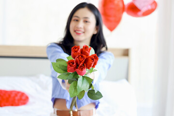 Beautiful red rose bouquet on Asian woman hand, happy girl showing Valentine flower present from lover boyfriend to camera while sitting on white bed in bedroom. Happy Saint Valentine Day.