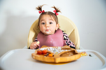 Portrait of cute toddler caucasian brunette girl with two tails eating by herself from modern wooden dish with a fork; self feeding concept, blw