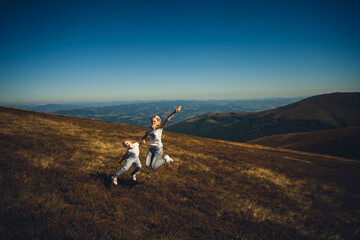 Obraz premium Happy family, mother and daughter, in summer clothes, having fun and laughing, holding hands and pulling each other on top of a mountain, on a sunny day in summer.