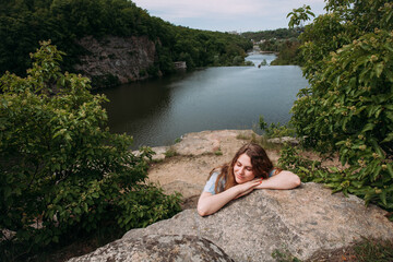 A young slender girl in a blue sundress, sits on the rocks near the rocks, against the backdrop of a picturesque river, rests and smiles, on a bright sunny day.