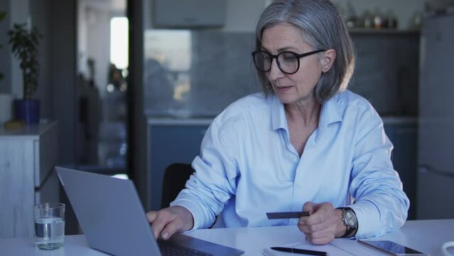 Mature woman in big eyeglasses doing shopping online, entering credit card number into web-site on laptop, choosing goods and paying for home delivery