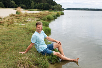 Naklejka na ściany i meble A smiling man with an athletic body, dressed in denim shorts and a blue t-shirt, sits on the banks of a picturesque river during the day.