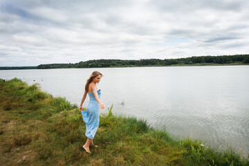 A beautiful slender smiling girl with long blond hair, in a blue summer dress, spins and walks along the river bank, against the backdrop of a picturesque landscape.