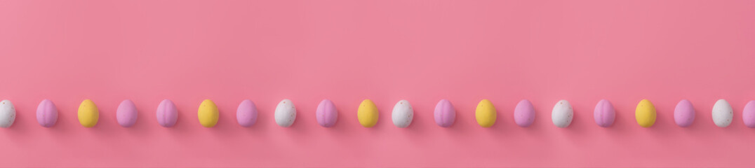 Easter banner made of sweet colorful small eggs lying in one line on pale pink background. Festive sweets. Copy space for text. Lying flat. Postcard.