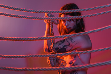 Tattooed model holding rope and looking at camera on purple background with light.