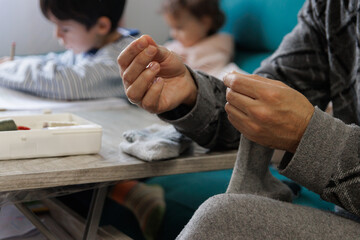 Fototapeta premium Man sewing a sock in the living room at home while his children do their homework
