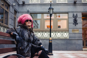 Fototapeta na wymiar Fashionable pretty woman in sunglasses wearing natural fur coat and pink fur hat sitting on bench in city district, looking away. Stylish lady posing outdoor. Fashion style concept. Copy space for ad