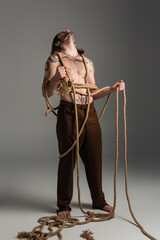 Log haired and tattooed model in pants holding rope on grey background.