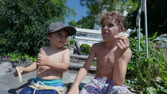 Two Little Brothers Hanging Out Together Outside. Younger Brother Nex To Older Sibling During Summer Holidays In The Sunlight Shirtless. Kids Friends Eating Snack