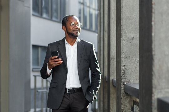 Portrait of a smiling confident and successful African American businessman in a suit and glasses standing outside near an office center, holding a phone in his hand. He keeps his hand in his pocket,