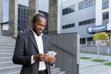 Happy African-American man, office worker, businessman stands near office center, holds cash in hands, counts money. Received financial reward, salary, bonus.