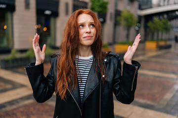 Portrait of frustrated red-haired young woman touching wet hair after autumn rain standing on beautiful city street. Front view of upset female untangling hair after being caught in rain.