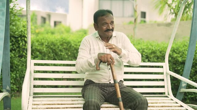 Indian Hindu lonely sad upset old aged senior elder widower man feel bad living with disability chronic health issue sitting on garden swing hold stick cane worried facing problem senility concept