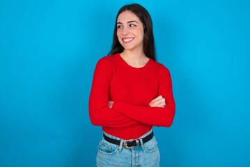 Dreamy rest relaxed young brunette girl wearing red T-shirt against blue wall crossing arms, looks good copyspace
