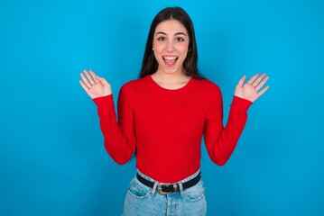 Optimistic young brunette girl wearing red T-shirt against blue wall raises palms from joy, happy to receive awesome present from someone, shouts loudly,