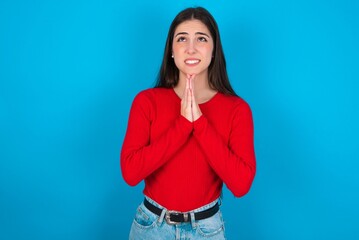 young brunette girl wearing red T-shirt against blue wall begging and praying with hands together with hope expression on face very emotional and worried. Please God
